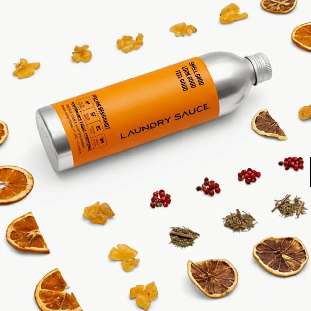 A silver bottle of laundry sauce with a bright orange label lies diagonally on a white background, surrounded by scattered dried orange slices, small yellow flower pieces, red berries, and herbs. The bottle label reads 'LAUNDRY SAUCE' with additional text including 'FRESH BERRY' and 'ORANGE'.