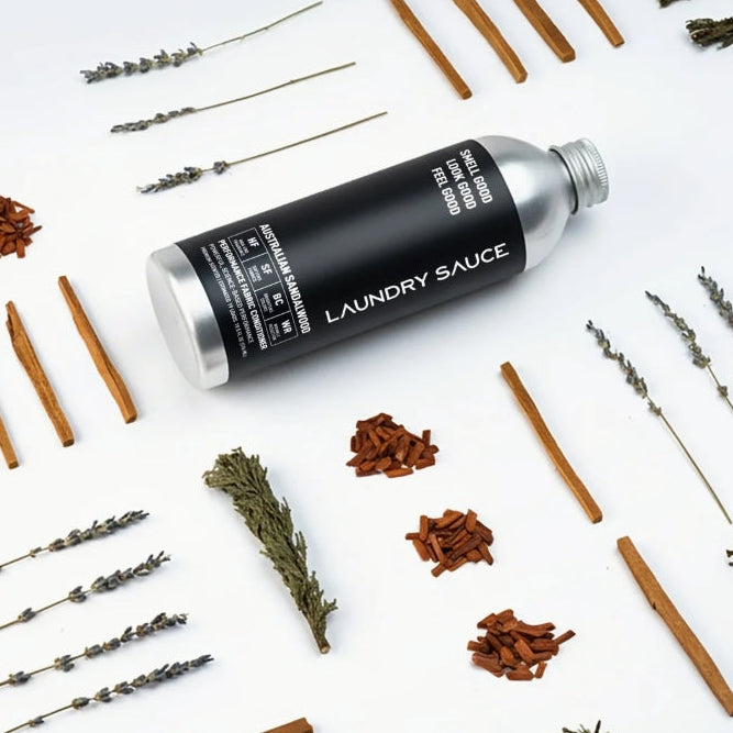 A black cylindrical laundry sauce bottle with a silver cap, surrounded by dried lavender sprigs, cinnamon sticks, and cloves on a white background.