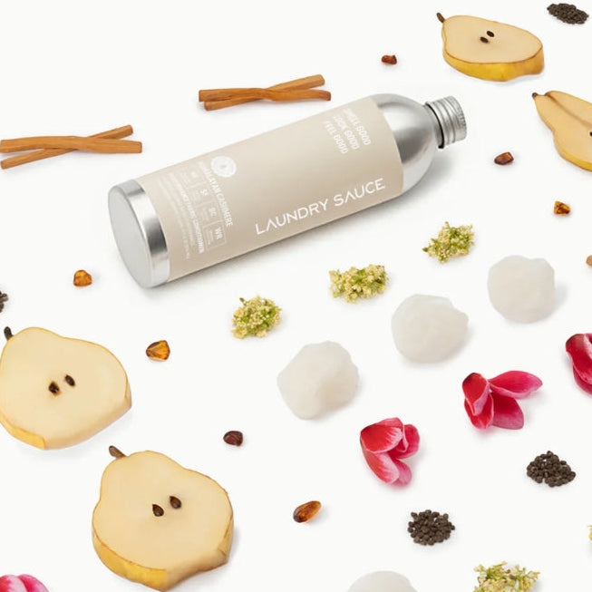 A silver and beige cylindrical bottle labeled 'LAUNDRY SAUCE' lies diagonally on a white background, surrounded by styled botanical props including sliced pears, cinnamon sticks, flower petals, and dried botanical pieces.