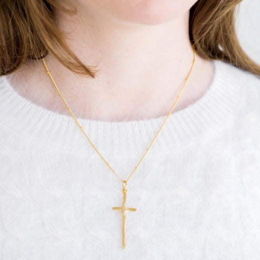 A close-up of a person wearing a delicate gold cross pendant necklace over a white textured knit sweater.