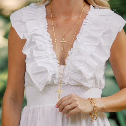 A woman wearing a white ruffled sleeveless top and light skirt, styled with layered gold necklaces featuring cross pendants and a gold chain bracelet, set against a soft green outdoor background.