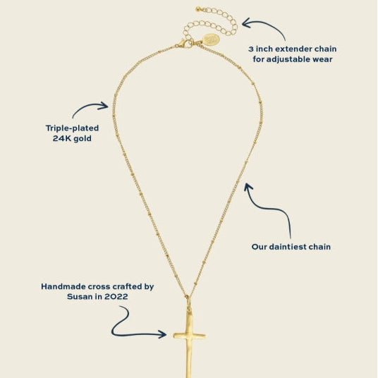 A gold cross pendant necklace with an adjustable chain, shown on a light beige background with descriptive callout text and arrows pointing to features like the triple-plated 24k gold chain, 3-inch extender chain, and handmade cross crafted by Susan in 2022.