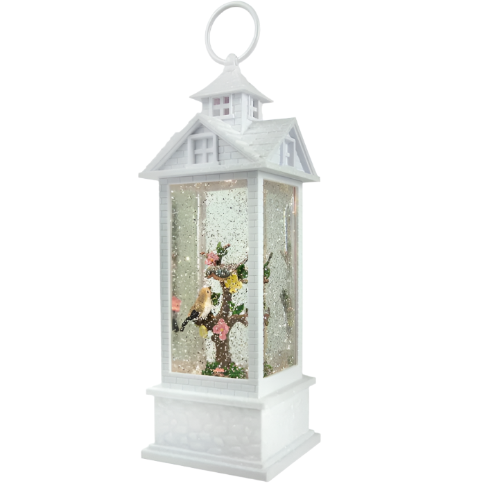 A decorative tabletop water gazebo lantern featuring white birds, a tree with pink and yellow flowers, and a colonial house exterior. The lantern is illuminated from the inside with glitter and lights.