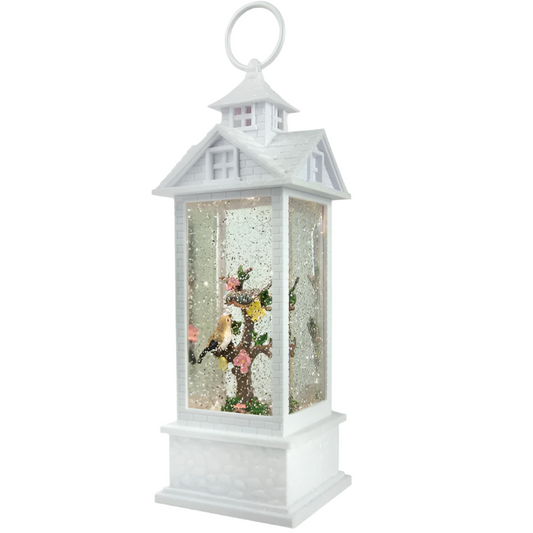 A decorative tabletop water gazebo lantern featuring white birds, a tree with pink and yellow flowers, and a colonial house exterior. The lantern is illuminated from the inside with glitter and lights.