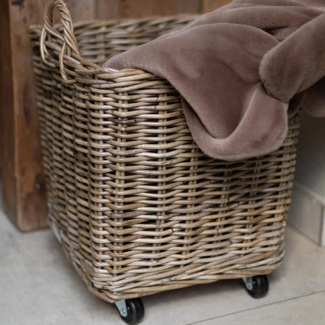 A natural wicker laundry hamper with black caster wheels, filled with a dusty taupe-brown plush blanket, set in a home environment next to a rustic wooden cabinet.