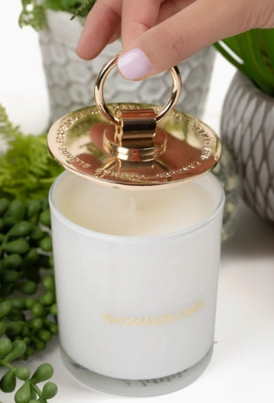 A white glass jar candle with a metallic rose gold ring handle lid, being lifted by a hand. The lid features gold lettering that reads 'THOMAS BURGE'.