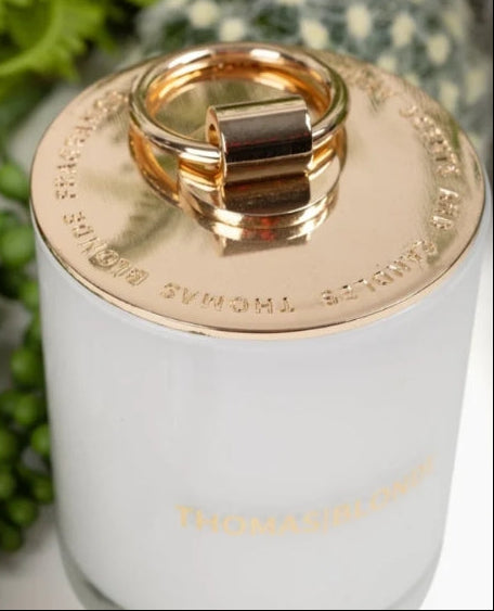 A close-up of a Thomas Sびlborne reed diffuser with a white glass body, a gold metal cap, and a gold ring handle, set against a soft-focus background with green foliage.