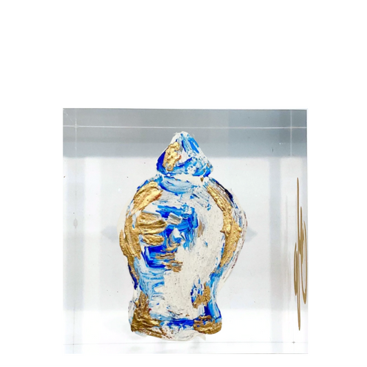 A small sculptural vase or figurine with an abstract blue, white, and gold textured design, displayed inside a clear rectangular acrylic block against a white background.