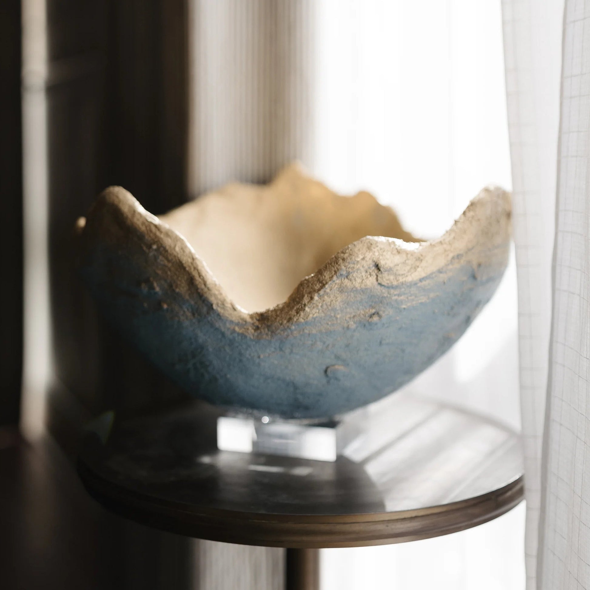 A rustic ceramic bowl with a textured, hand-painted blue exterior and a natural beige interior, displayed on a small round table in front of a window with sheer curtains.