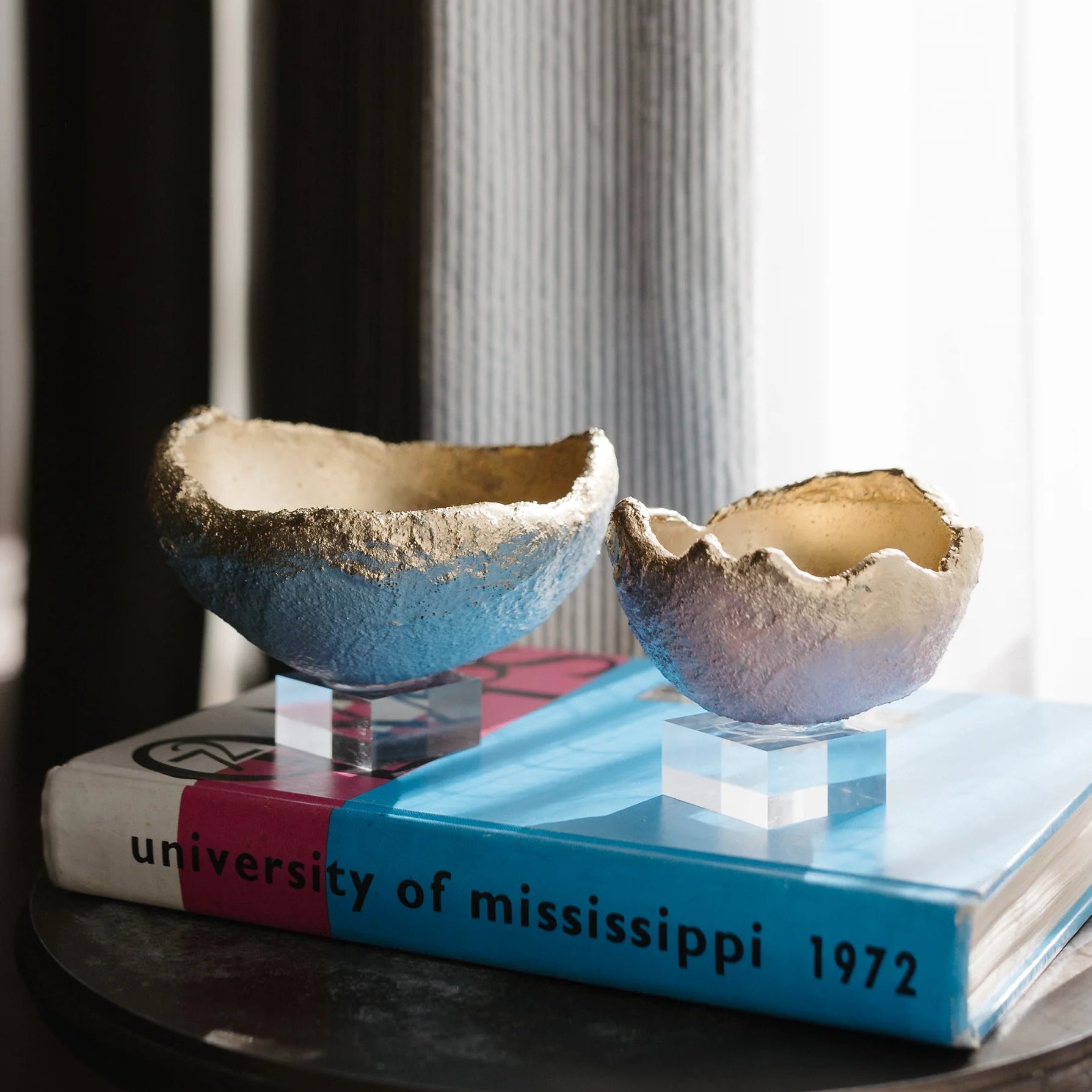 Two handmade ceramic egg-shaped bowls with a blue and beige glaze sit on a stack of books on a small round table in front of a window with sheer curtains.