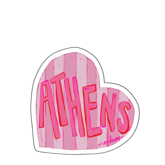 A die-cut heart-shaped sticker featuring the word 'ATHENS' in stylized pink lettering over a pink and white striped background, with a small signature in the lower right corner, shown on a plain white background.