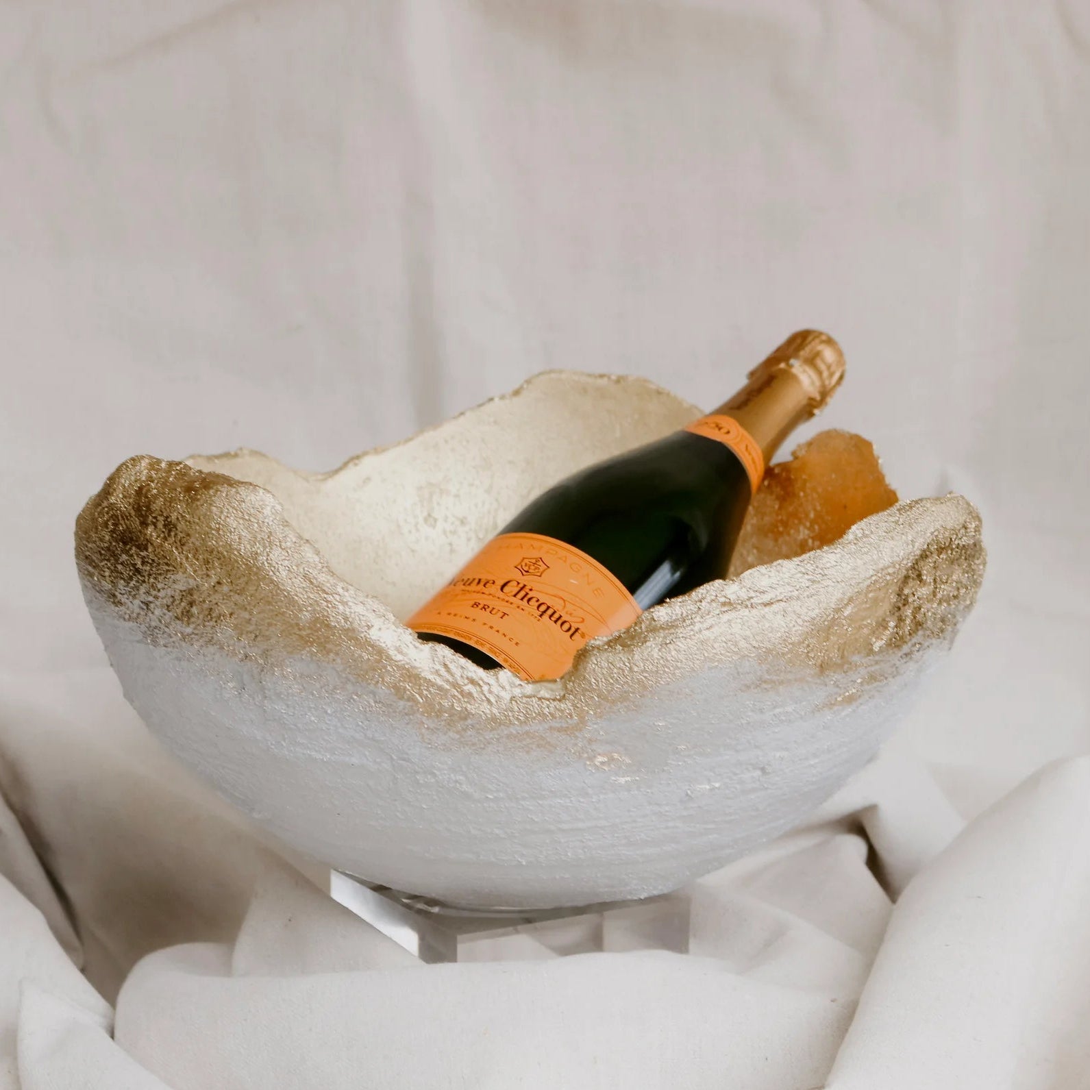 A textured white stone wine bowl with a gold rim, holding a bottle of Veuve Clicquot champagne on a clear stand, set against a draped white fabric background.