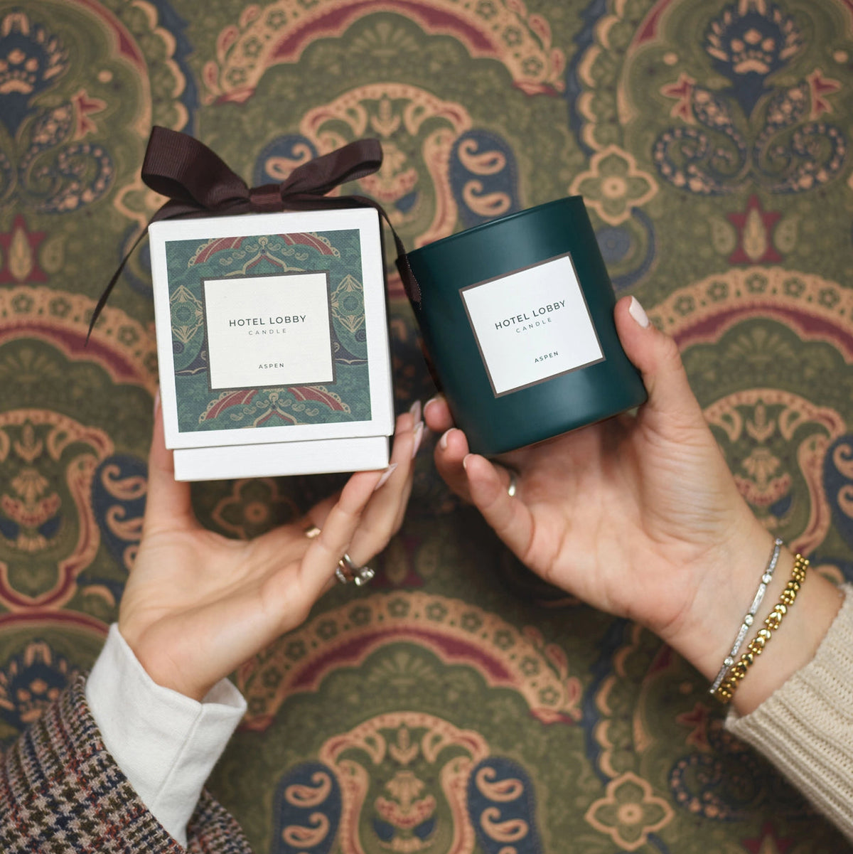 Two hands hold a candle and its gift box against a detailed paisley patterned fabric background. The candle is in a dark teal-green glass jar with a white label reading 'HOTEL LOBBY CANDLE', and the box is white with a dark brown ribbon and matching label.