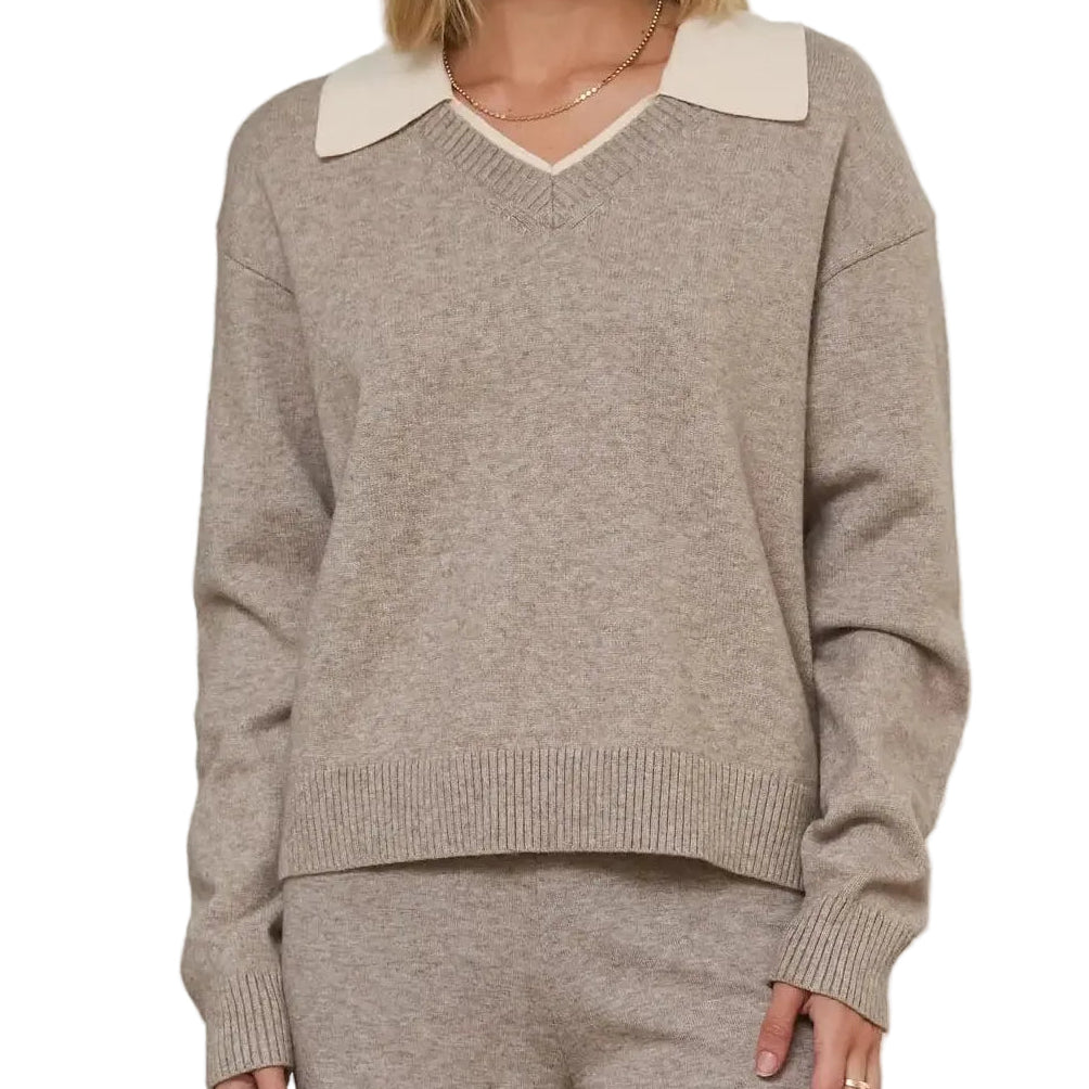 A woman modeling a taupe knit sweater with a contrasting cream V-neck collar, paired with matching taupe knit pants, set against a soft, neutral indoor background with textured columns.