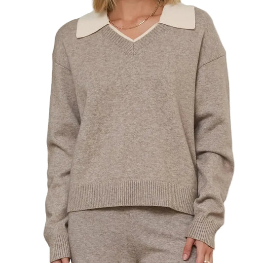 A woman modeling a taupe knit sweater with a contrasting cream V-neck collar, paired with matching taupe knit pants, set against a soft, neutral indoor background with textured columns.
