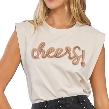 A woman modeling a sleeveless cream t-shirt with 'cheers!' in metallic rose gold lettering, paired with a black glittery mini skirt and black heels, standing in a bright white room with classical columns.